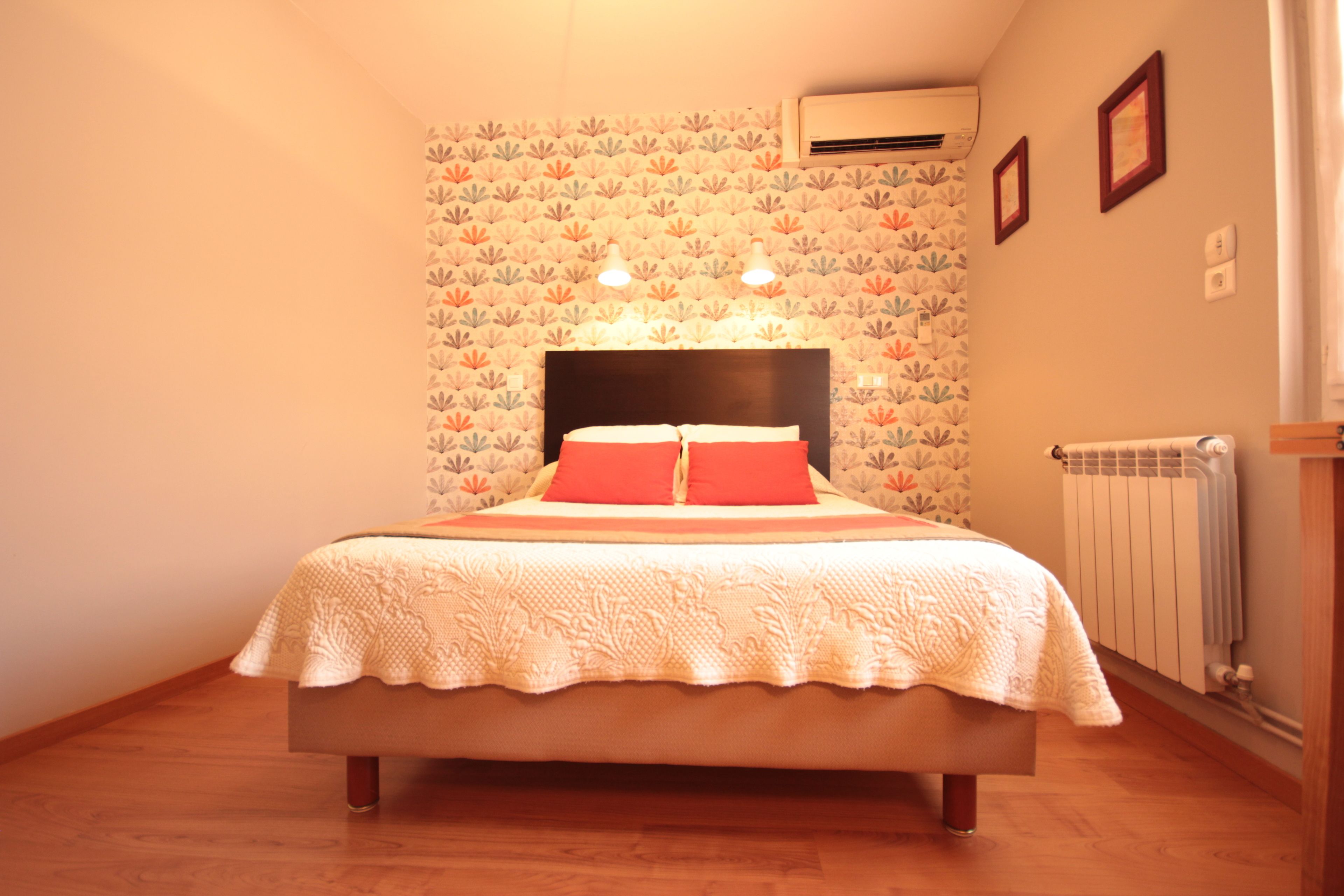 A bedroom with a bed, floral wallpaper, two wall-mounted lights, an air conditioner, and a radiator.