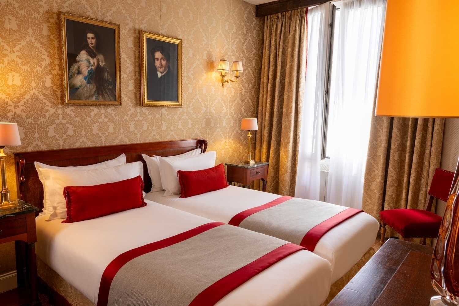 A hotel room with two beds, a wooden headboard, a nightstand with a lamp, a desk with a lamp, and a chair.