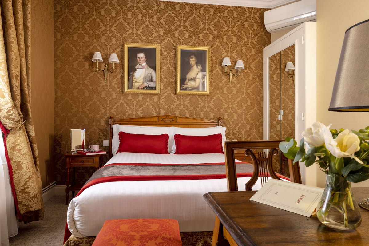Une chambre d’hôtel avec un lit, une table de chevet, un miroir, un bureau et un vase avec des fleurs.