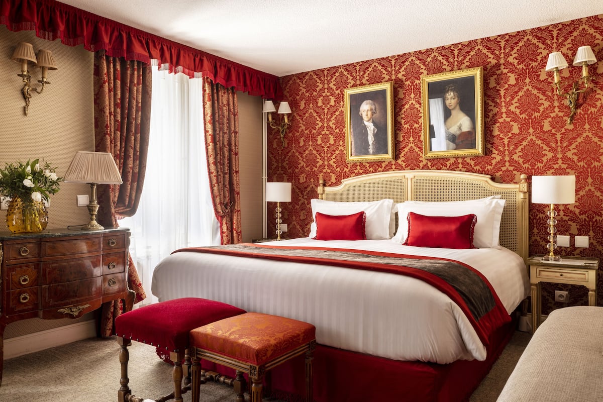 Une chambre d’hôtel classique, dotée d’un grand lit, d’un banc en bois, d’une commode et de deux lampes fixées au mur.