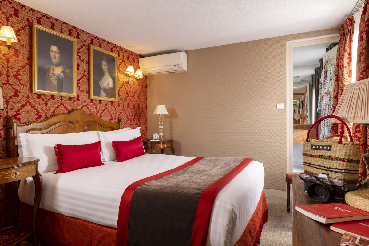 Une chambre d’hôtel avec un grand lit, deux tableaux accrochés au mur, un appareil de climatisation, une lampe de chevet, une table de chevet en bois et un panier en osier contenant un sac rouge.