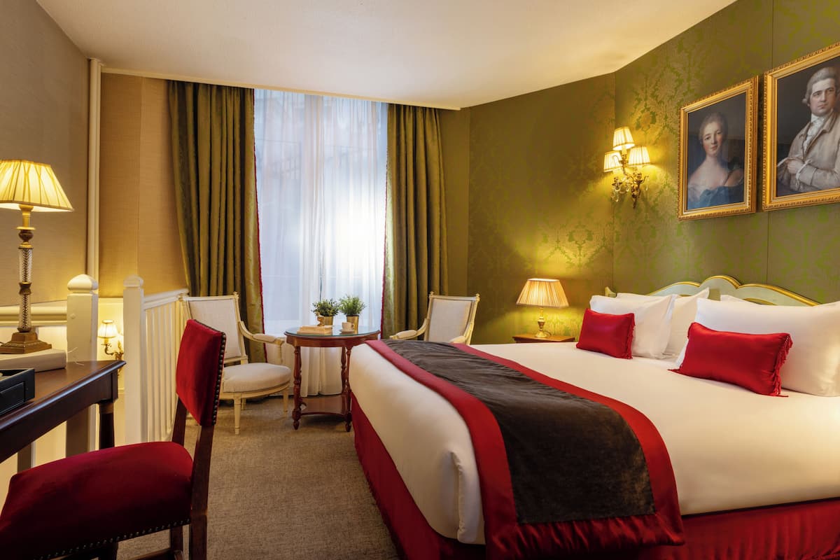 Une chambre d’hôtel avec un grand lit, deux chaises, une petite table avec une lampe et une fenêtre avec des rideaux.