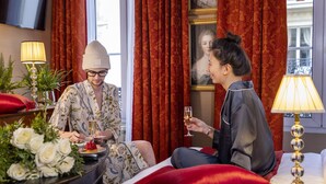 A hotel room with a bed, red curtains, a painting, and a table with a lamp and flowers. - Hotel de Seine (Paris)