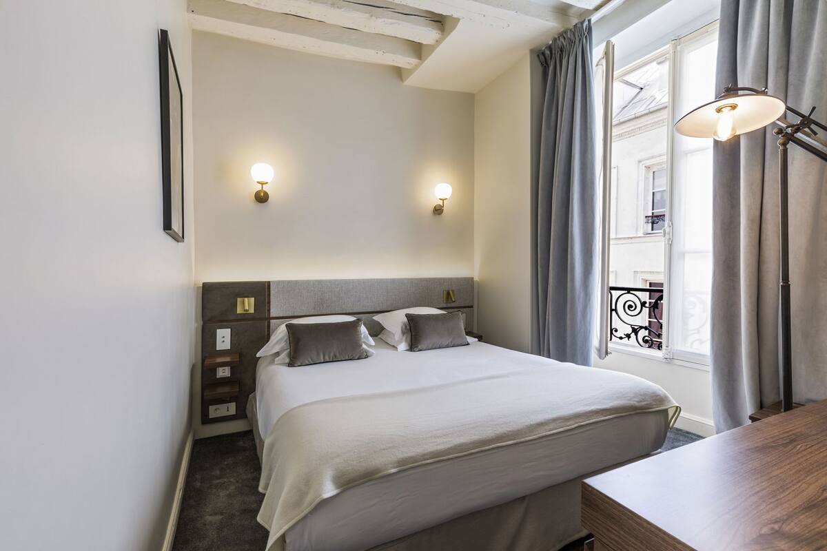 A hotel room with a bed, bedside tables, a desk, a window with curtains, and wall-mounted lights.