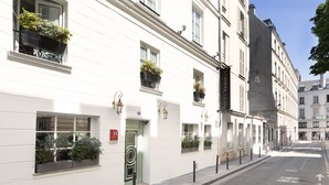 A white building with multiple windows, some with black wrought iron balconies and potted plants. There is a visible fire hydrant and a street with a clear sky. - Hotel Verneuil (Paris)