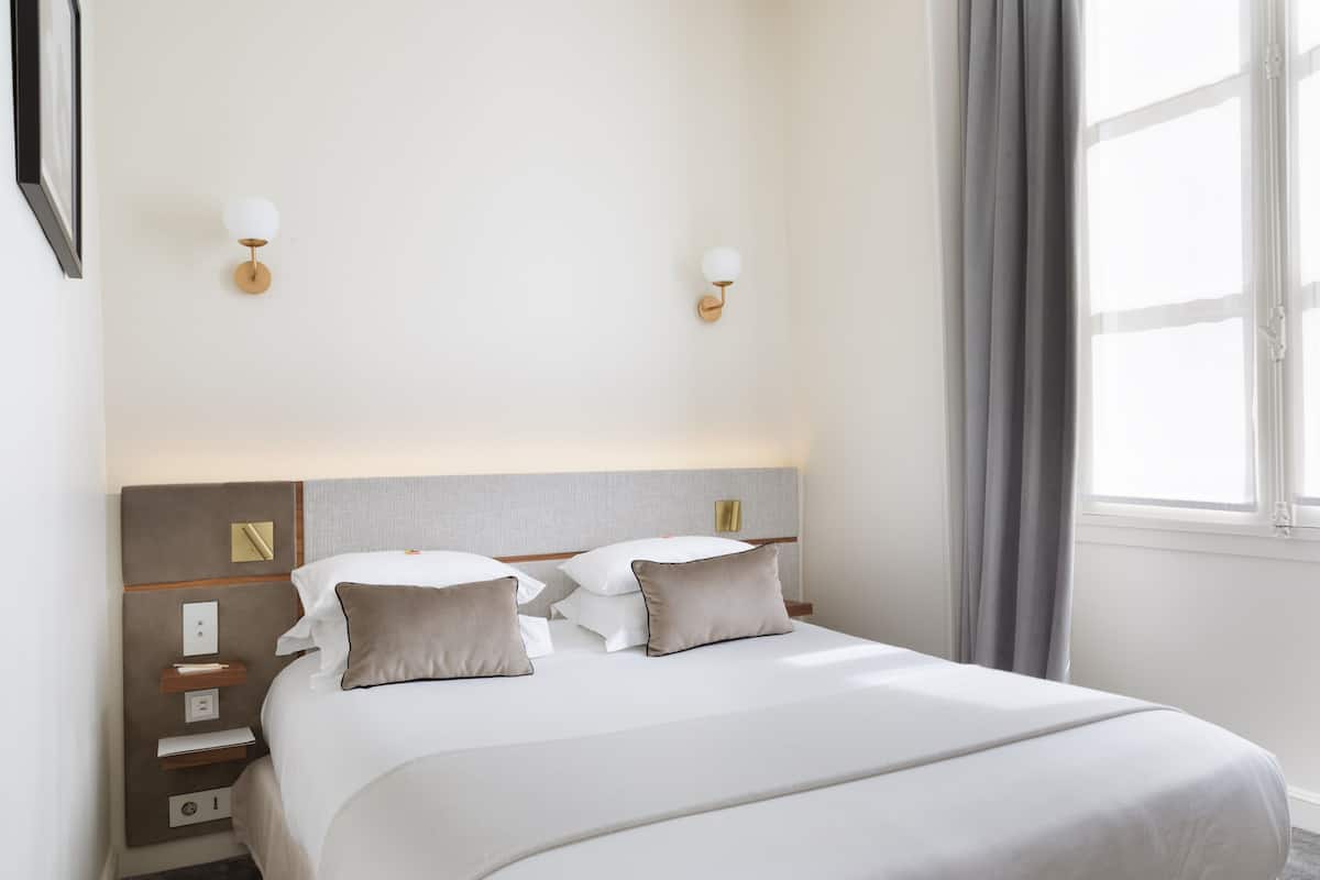A hotel room with a large bed, two bedside tables, a nightstand, a wall-mounted light fixture, and a window with curtains.