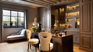 A cozy room with a sofa, a desk, and a chair. There is a window with curtains, a statue, and shelves with books and decorative items. - Hotel Verneuil (Paris)