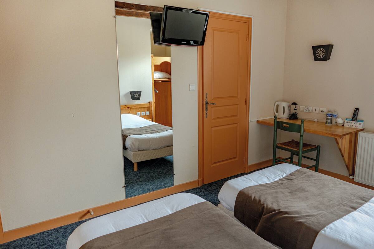 Ein Hotelzimmer mit zwei Betten, einem Schreibtisch mit Stuhl, einem an der Wand befestigten Fernseher und einer Tür zu einem weiteren Raum.