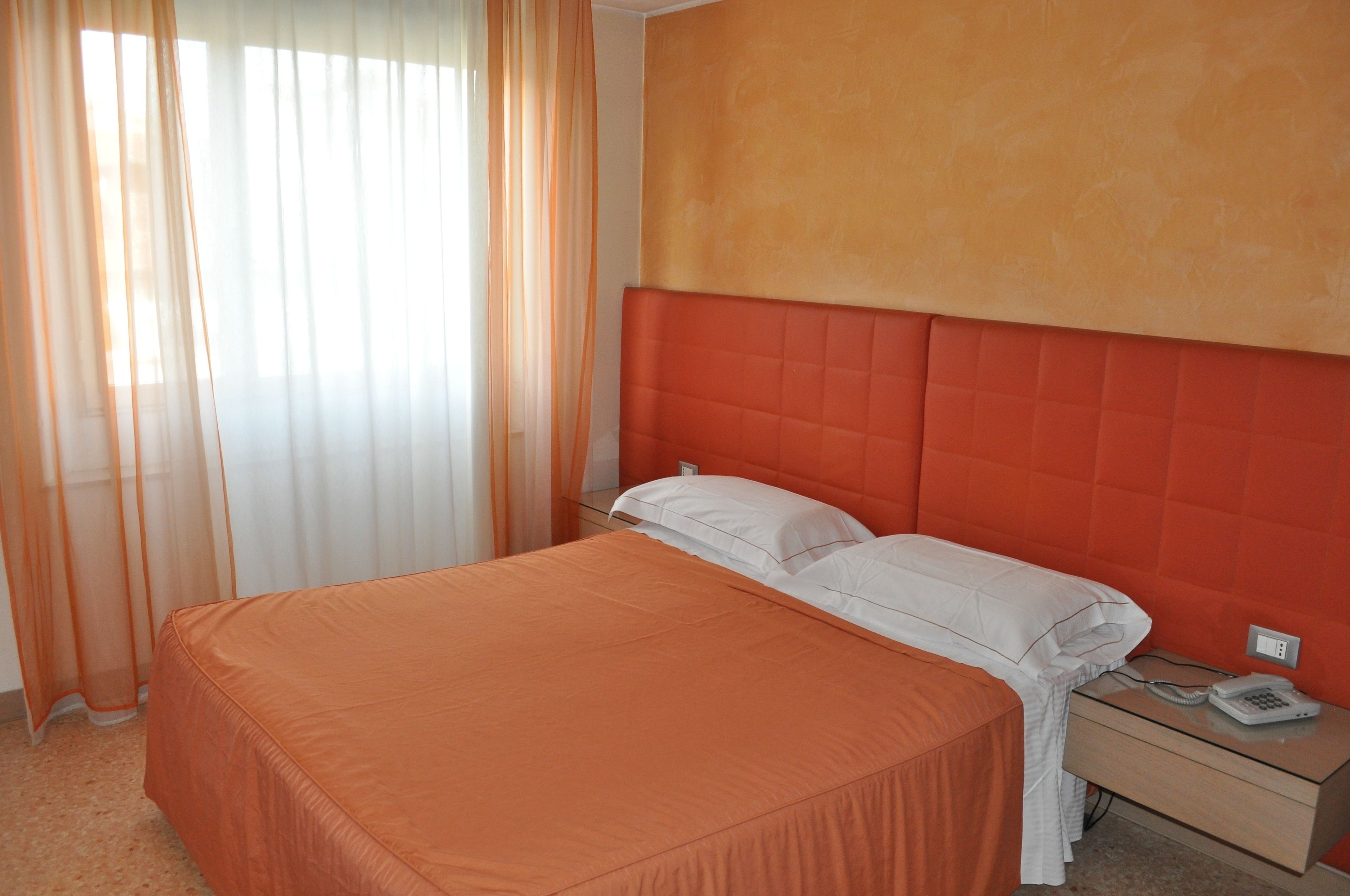 Una habitación de hotel con cama, mesita de noche, teléfono y cortinas translúcidas.