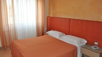 Una habitación de hotel con cama, mesita de noche, teléfono y cortinas translúcidas.