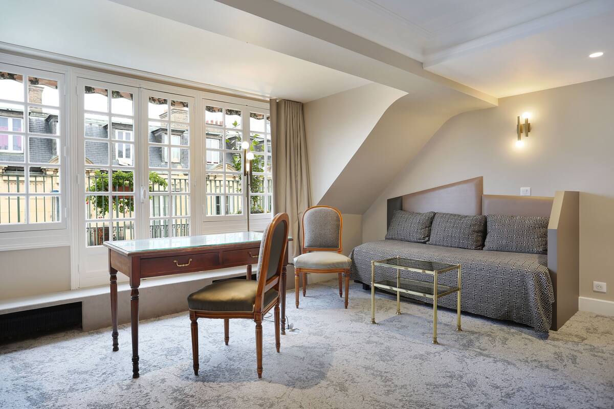 Ein Schlafzimmer mit Schreibtisch, zwei Stühlen, einem Bett und Blick auf Gebäude durch große Fenster.