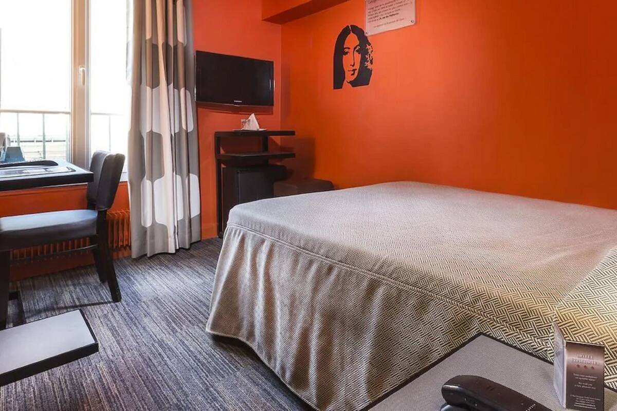 Ein Hotelzimmer mit einem Bett, einem Schreibtisch mit Computer, einem Sessel, einem an der Wand befestigten Fernseher und einem Fenster mit Vorhängen.