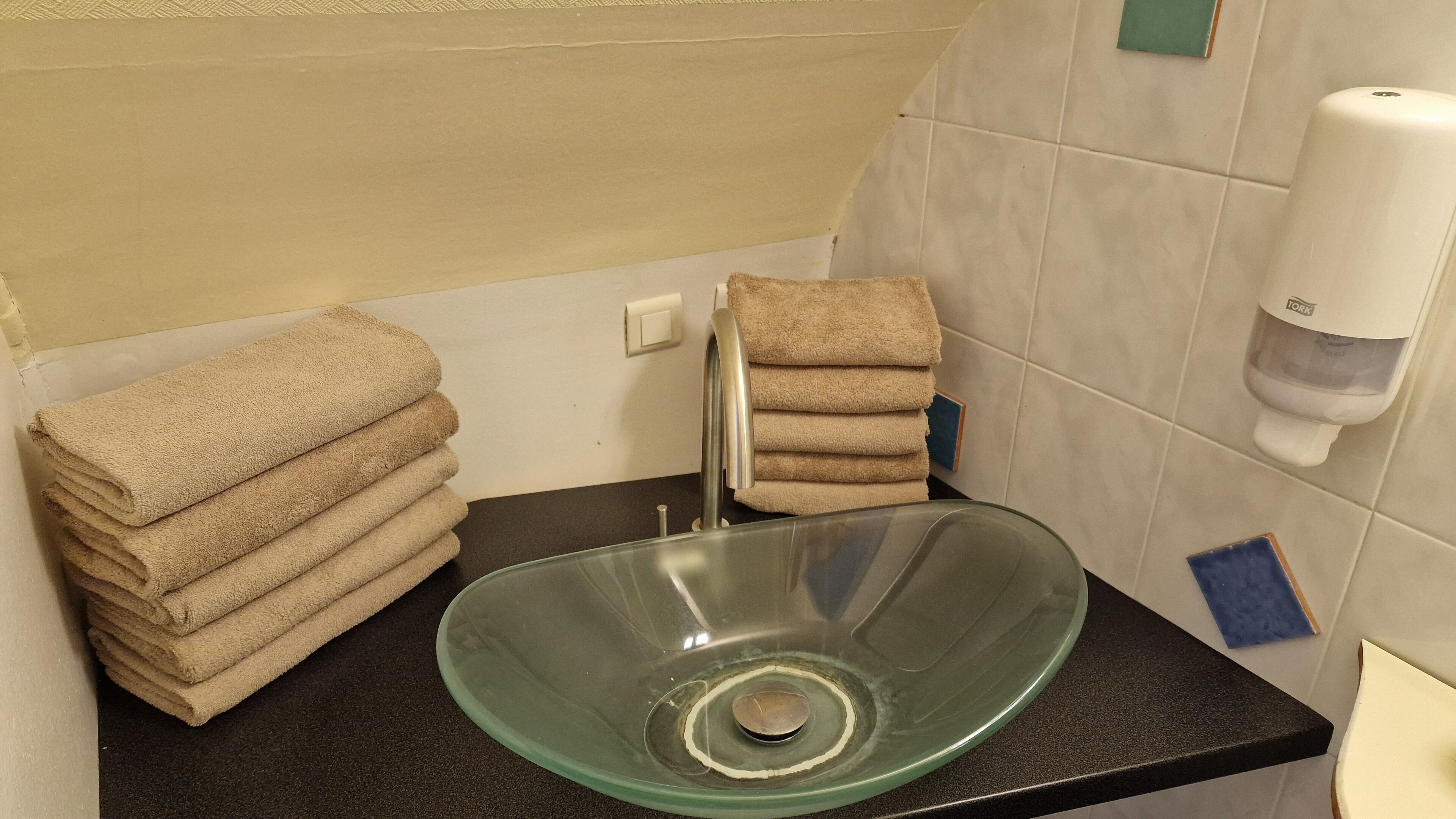 A bathroom with a glass sink, stacked towels, a soap dispenser, and a towel rack.