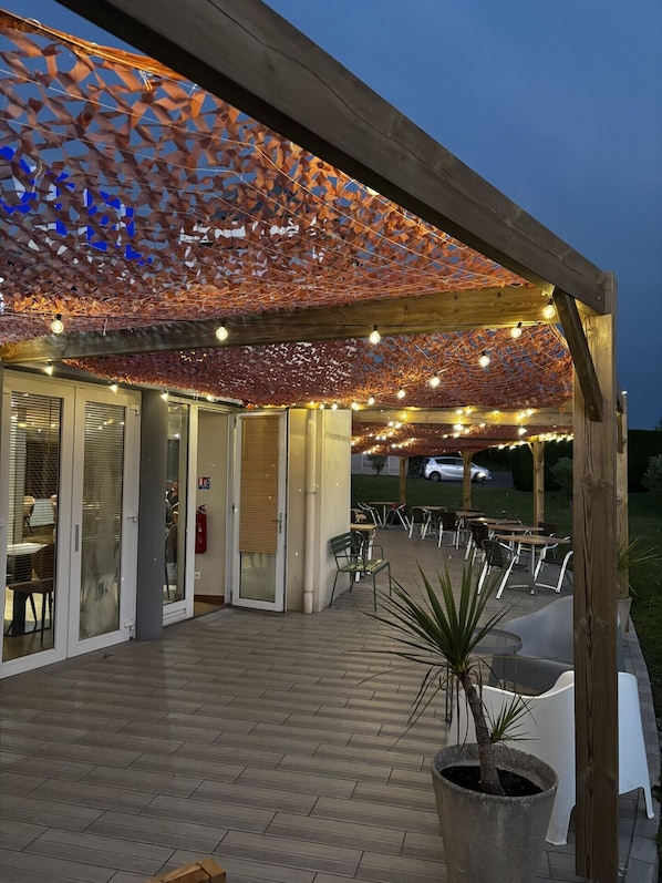 Un área de patio al aire libre con una pérgola cubierta de tela estampada, mesas y sillas dispuestas para comer, y una planta en maceta al frente.