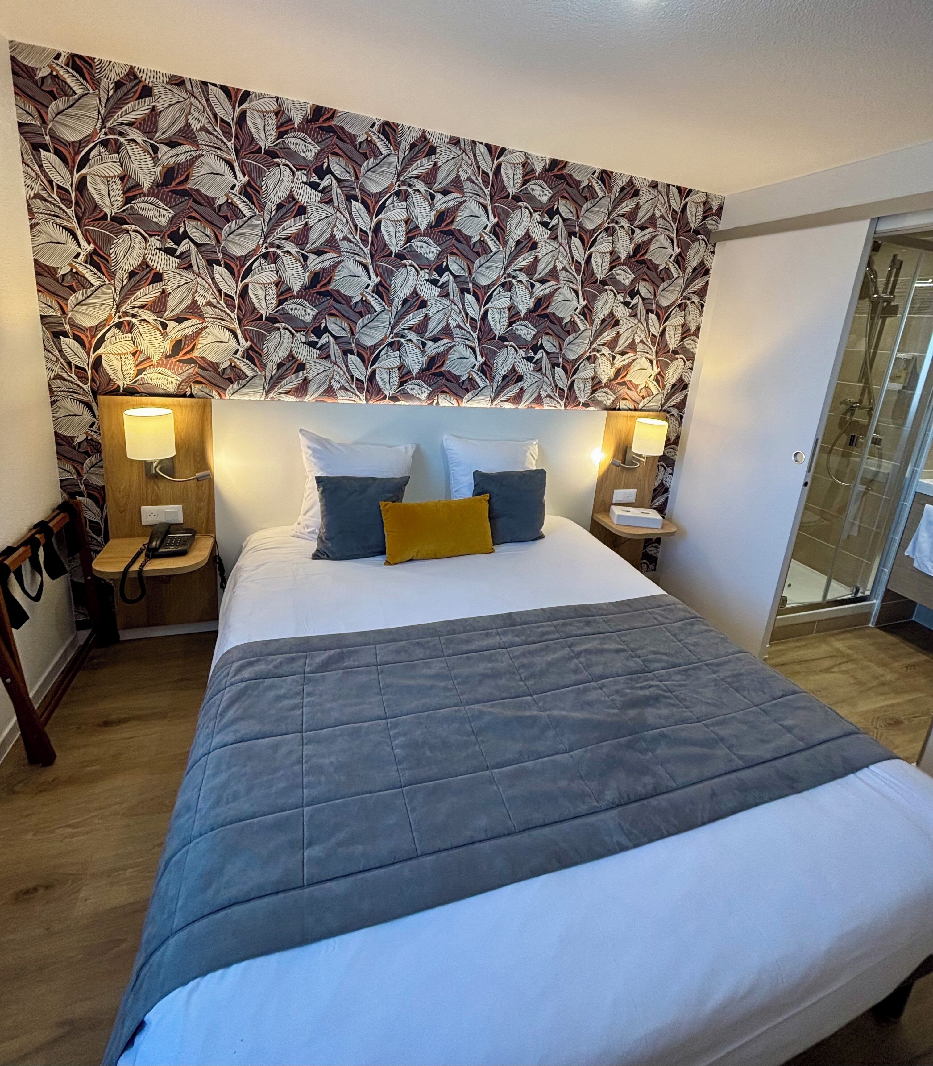A hotel room with a large bed, two bedside tables with lamps, a floral wallpaper headboard, and a glass shower door.