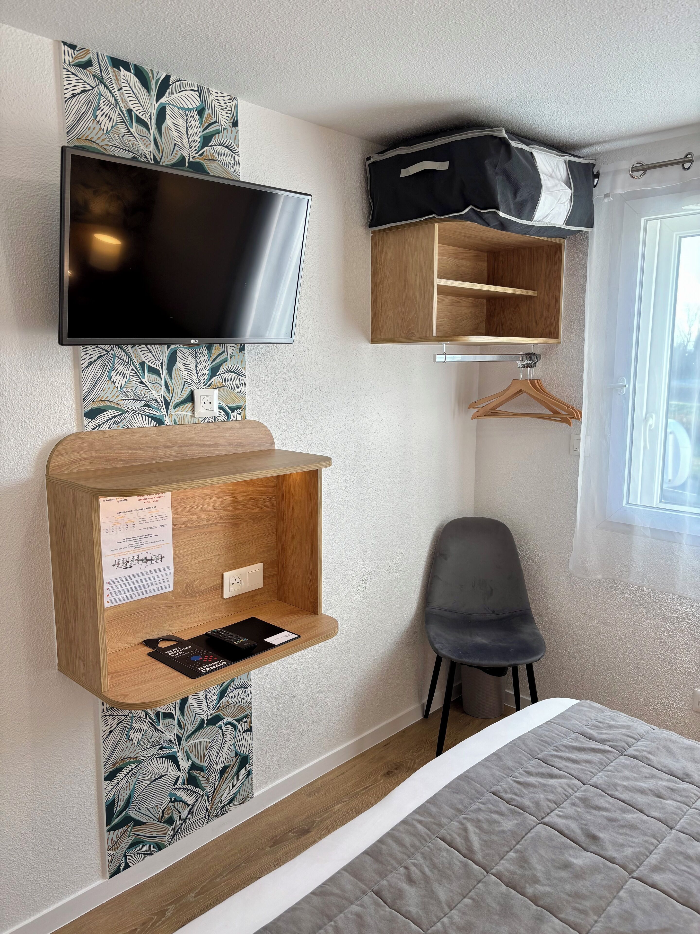 Habitación de hotel con televisor de pantalla plana montado en la pared, estante de madera con un escritorio pequeño, una silla gris y una cama con colcha gris.