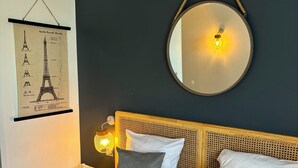 Una cama bien tendida con cabecera, un espejo redondo montado en la pared y un póster enmarcado de la Torre Eiffel.