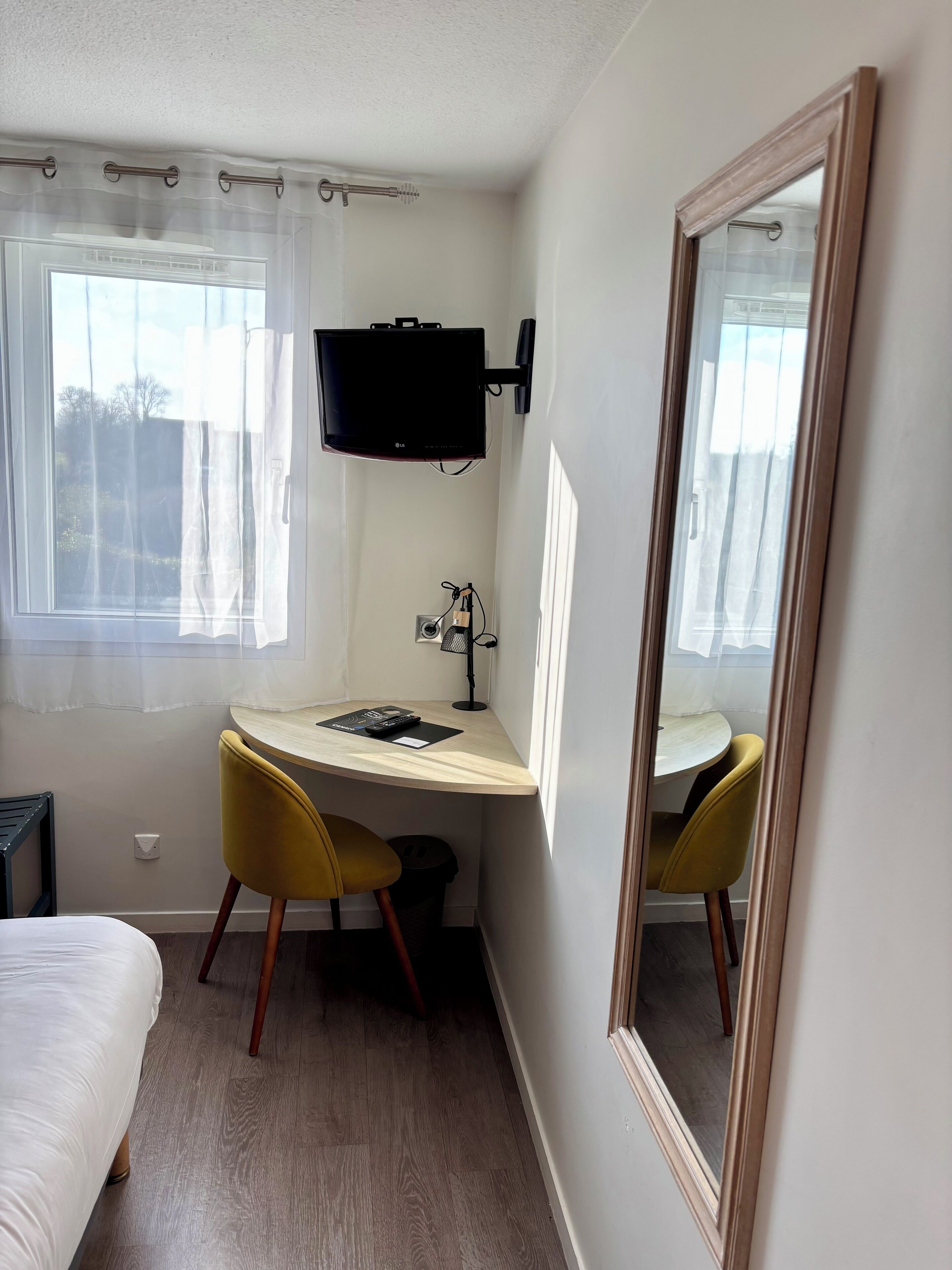 A small, well-lit room with a bed, a desk with a chair, a television mounted on the wall, and a large mirror.