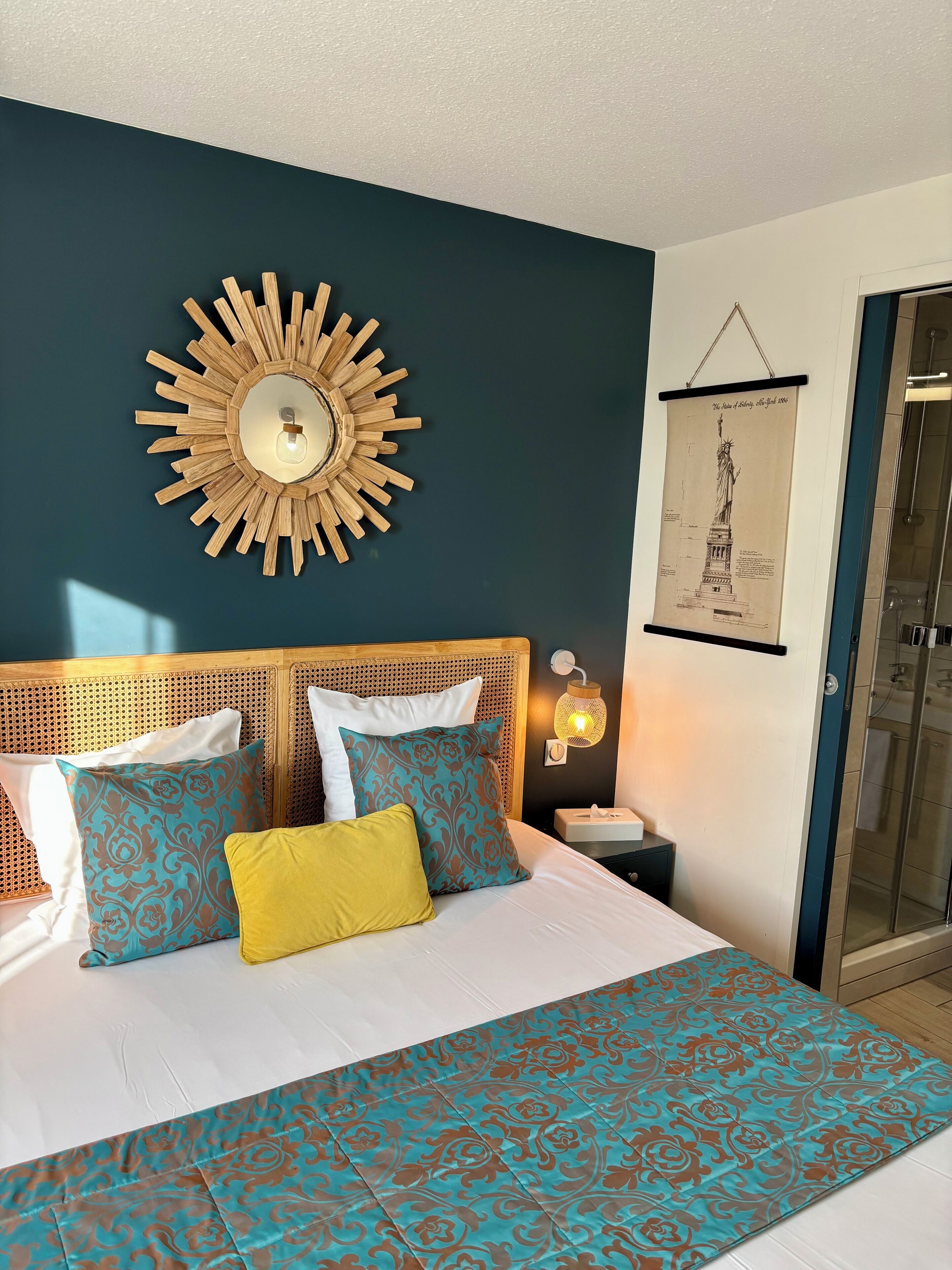 Une chambre à coucher avec un lit, une couvre-lit à motifs, un miroir décoratif fixé au mur et un tableau encadré représentant la Statue de la Liberté.