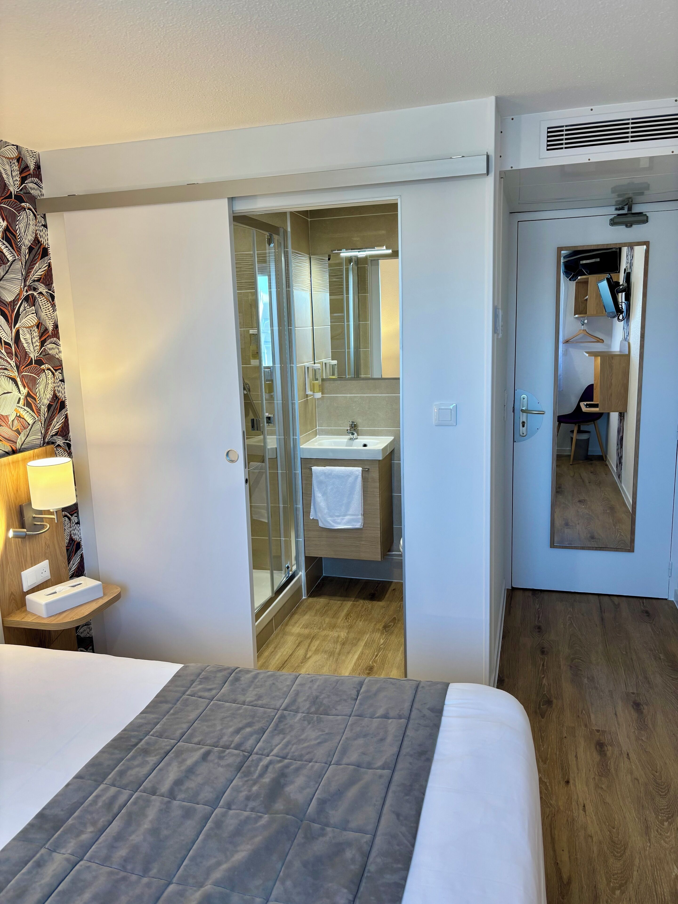 Habitación de hotel con cama, mesita de noche con lámpara, baño con lavamanos y espejo, y suelo de madera.