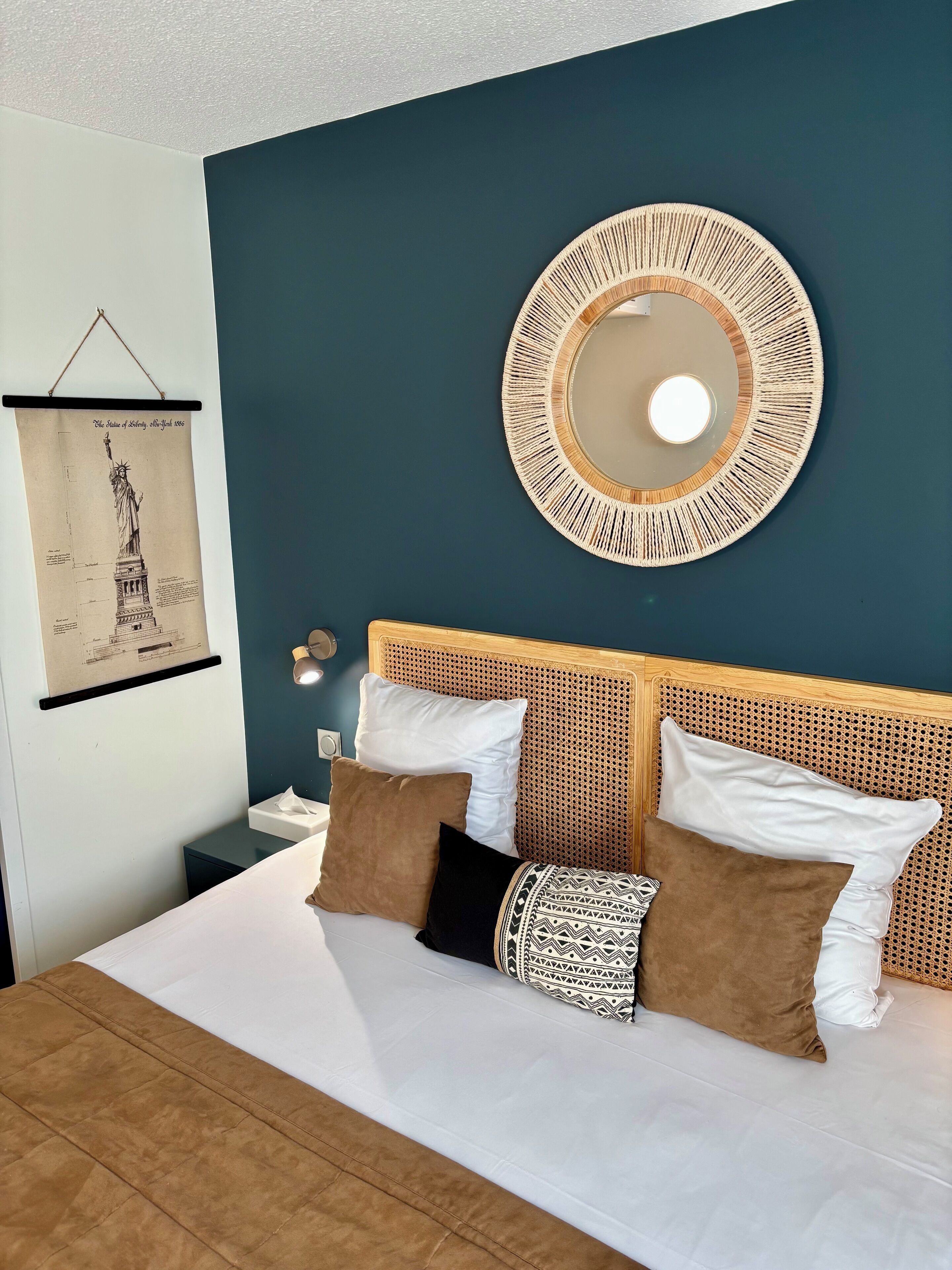 A neatly made bed with white and brown bedding, a round mirror with a woven frame, and a framed picture of the Statue of Liberty on the wall.