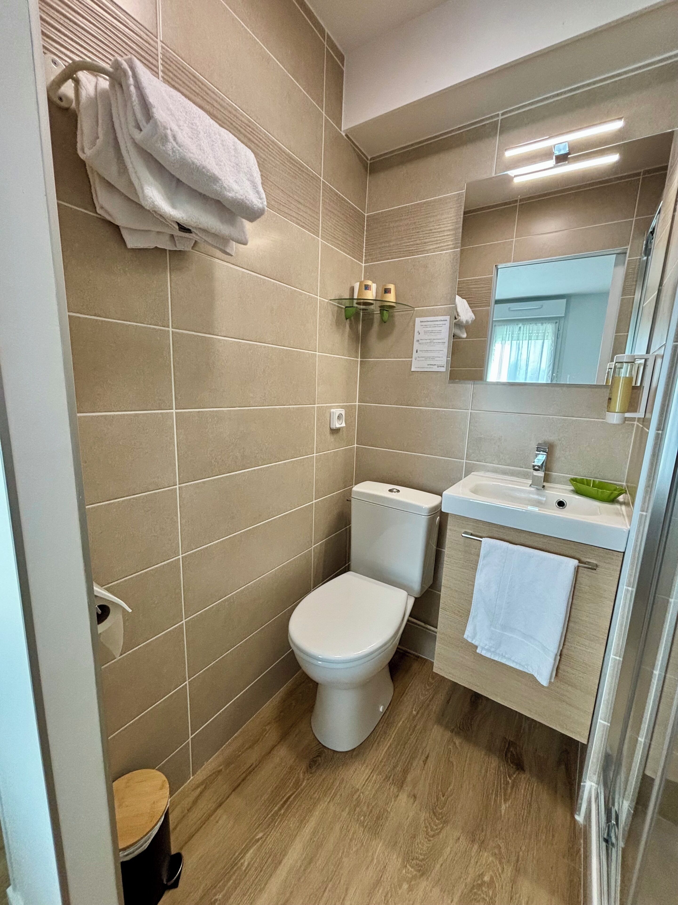 Un baño moderno con baldosas color beige, un inodoro blanco, un lavabo con un recipiente verde y un espejo.