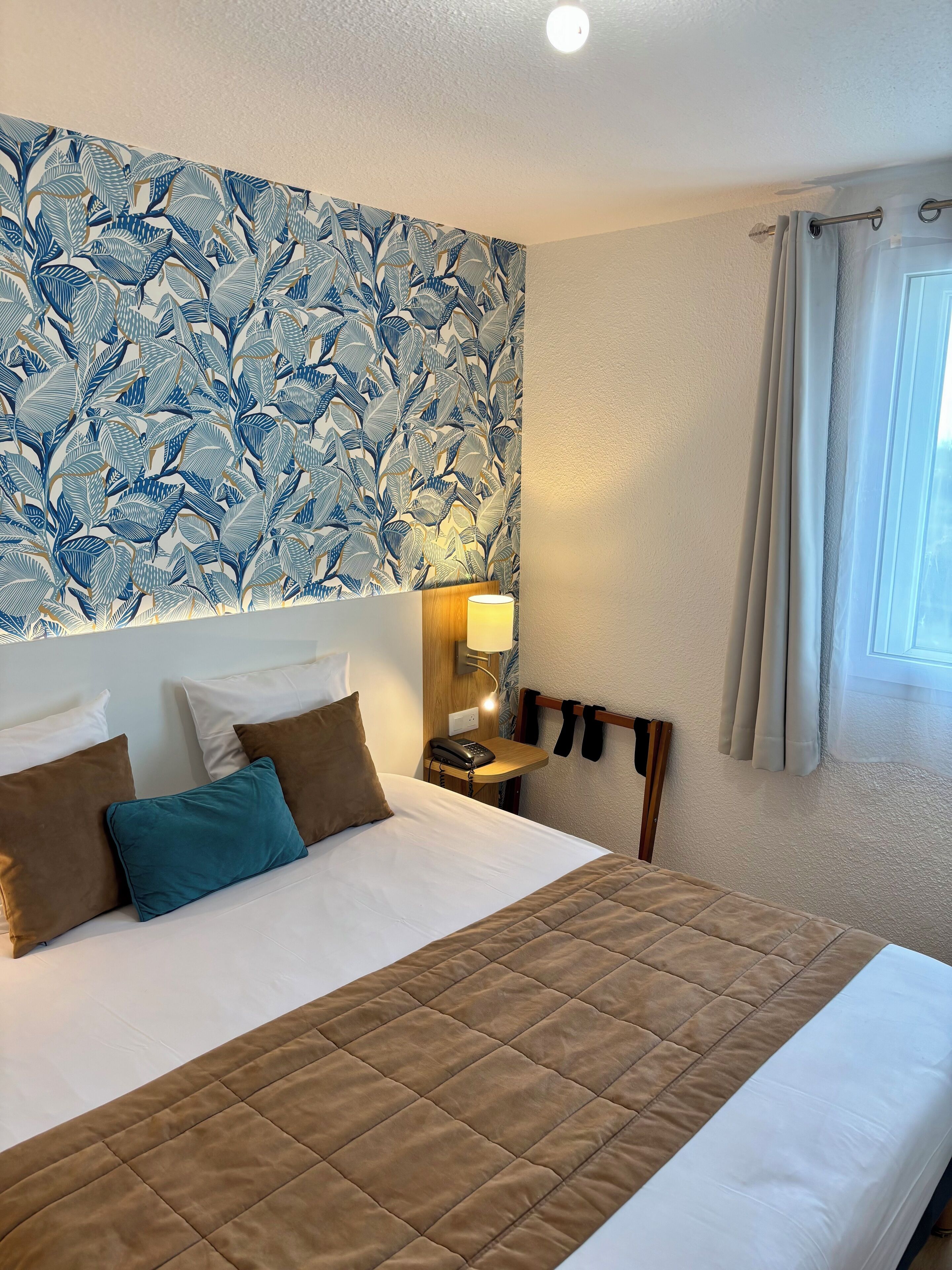 A hotel room with a bed, pillows, a bedside lamp, a telephone, and a window with curtains.