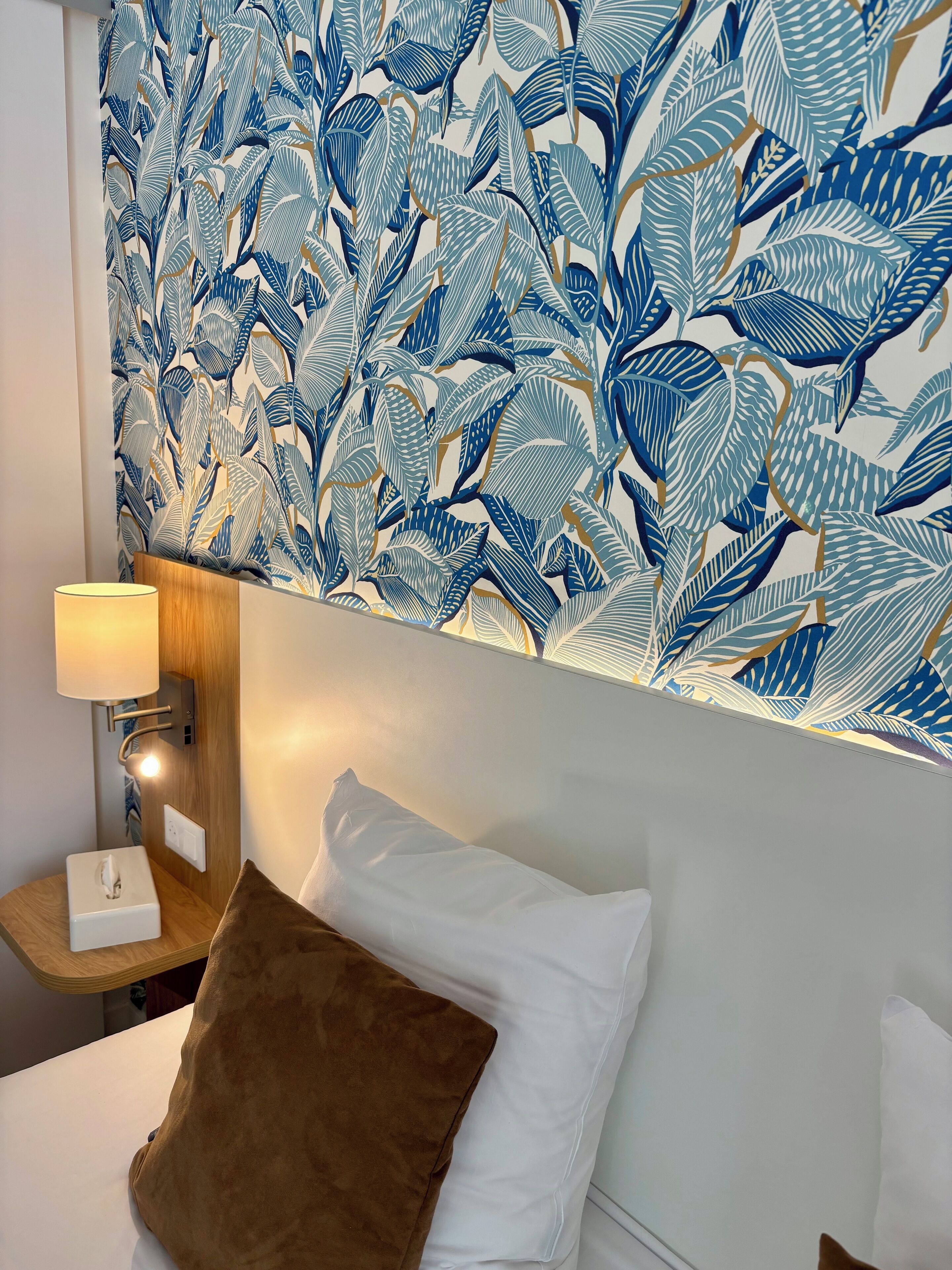 A hotel room with a bed, a nightstand, a lamp, and a wall with a leaf pattern.
