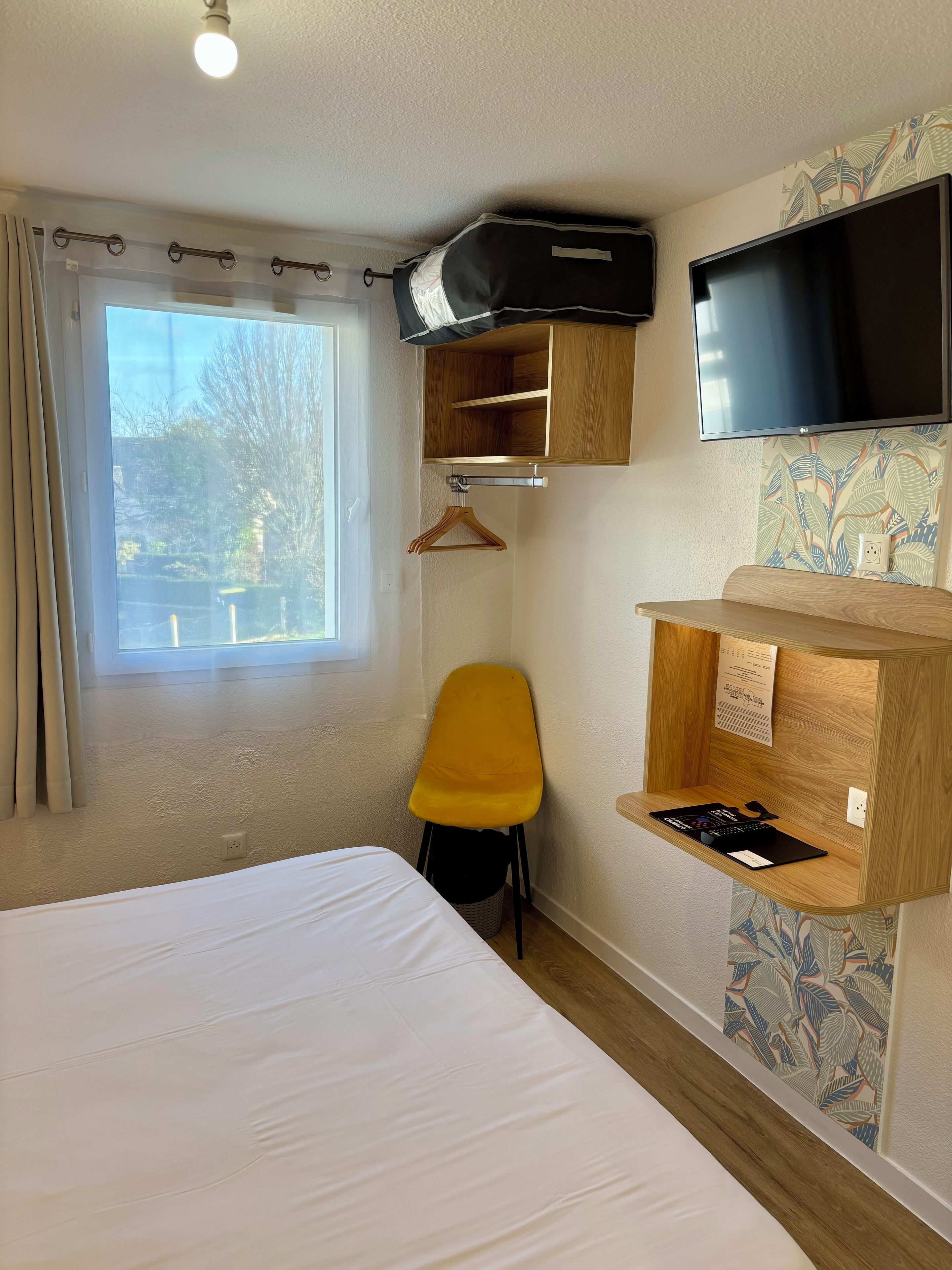 Une chambre d’hôtel avec un lit, une chaise jaune, un téléviseur fixé au mur et une étagère en bois avec une télécommande.
