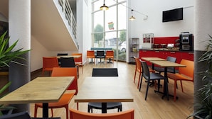 A modern café with wooden tables and orange chairs, large windows, and a red counter with a coffee machine. - hotelF1 Reims Tinqueux (Tinqueux)