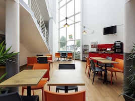 A modern café with wooden tables and orange chairs, large windows, and a red counter with a coffee machine.