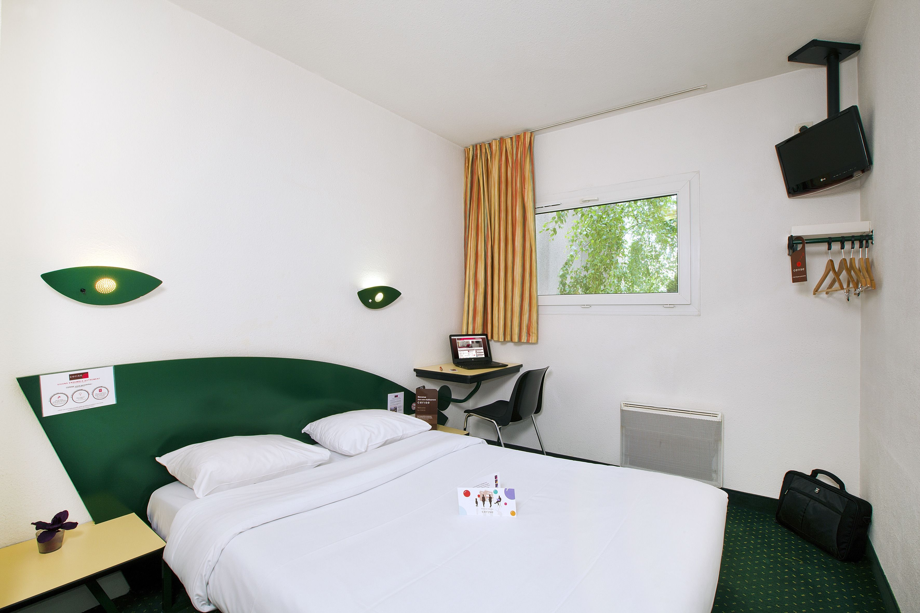 Una camera d'albergo con un letto, una scrivania, una sedia, una finestra e un termosifone.