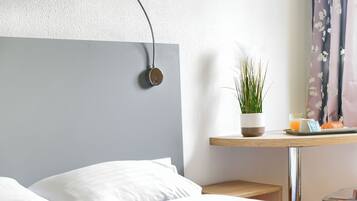 Una cama bien hecha con sábanas blancas, una mesita de noche con una planta en maceta, una lámpara y una bandeja pequeña con un croissant y un vaso de zumo de naranja.