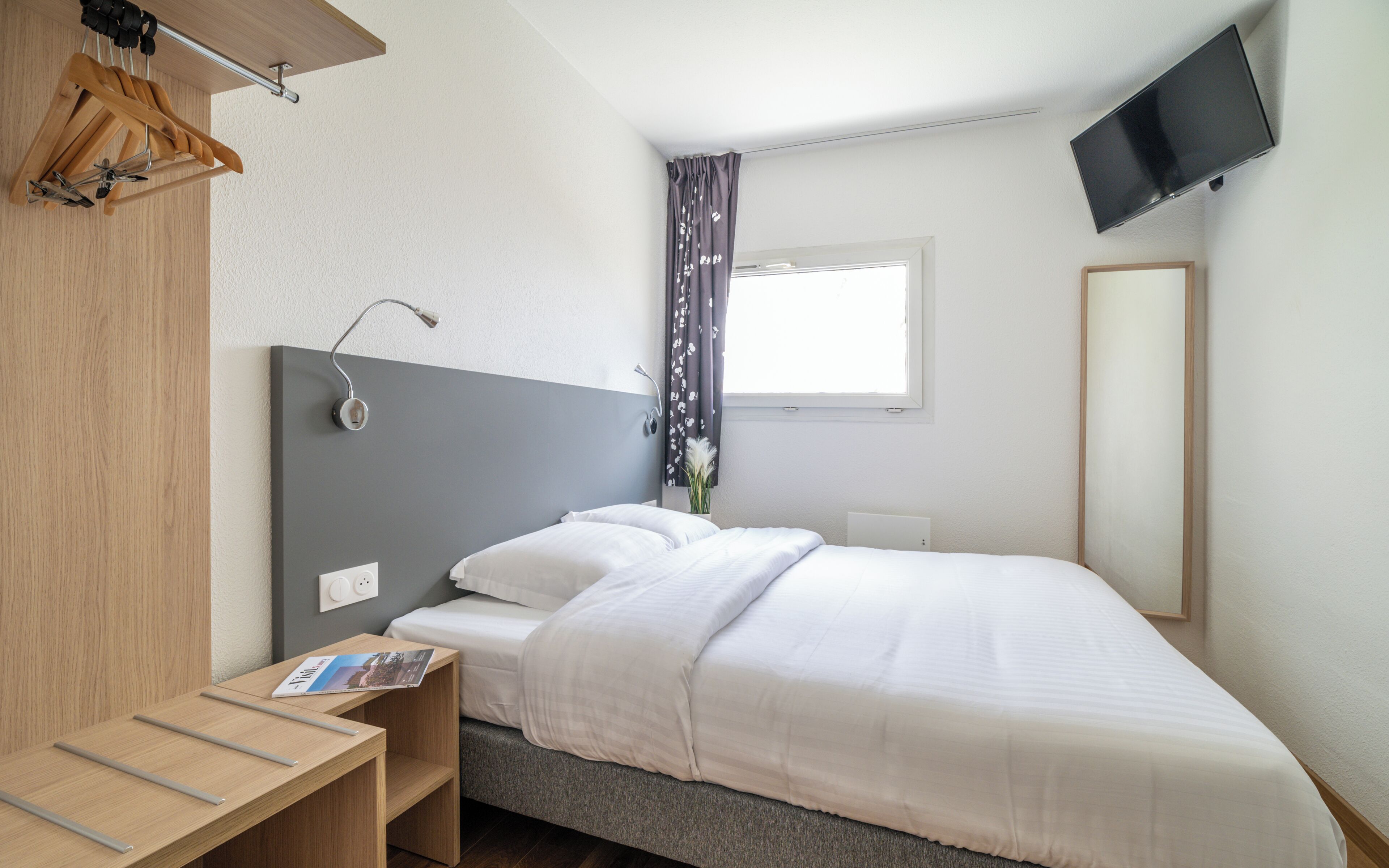 A hotel room with a bed, bedside table, lamp, TV, and a window with curtains.