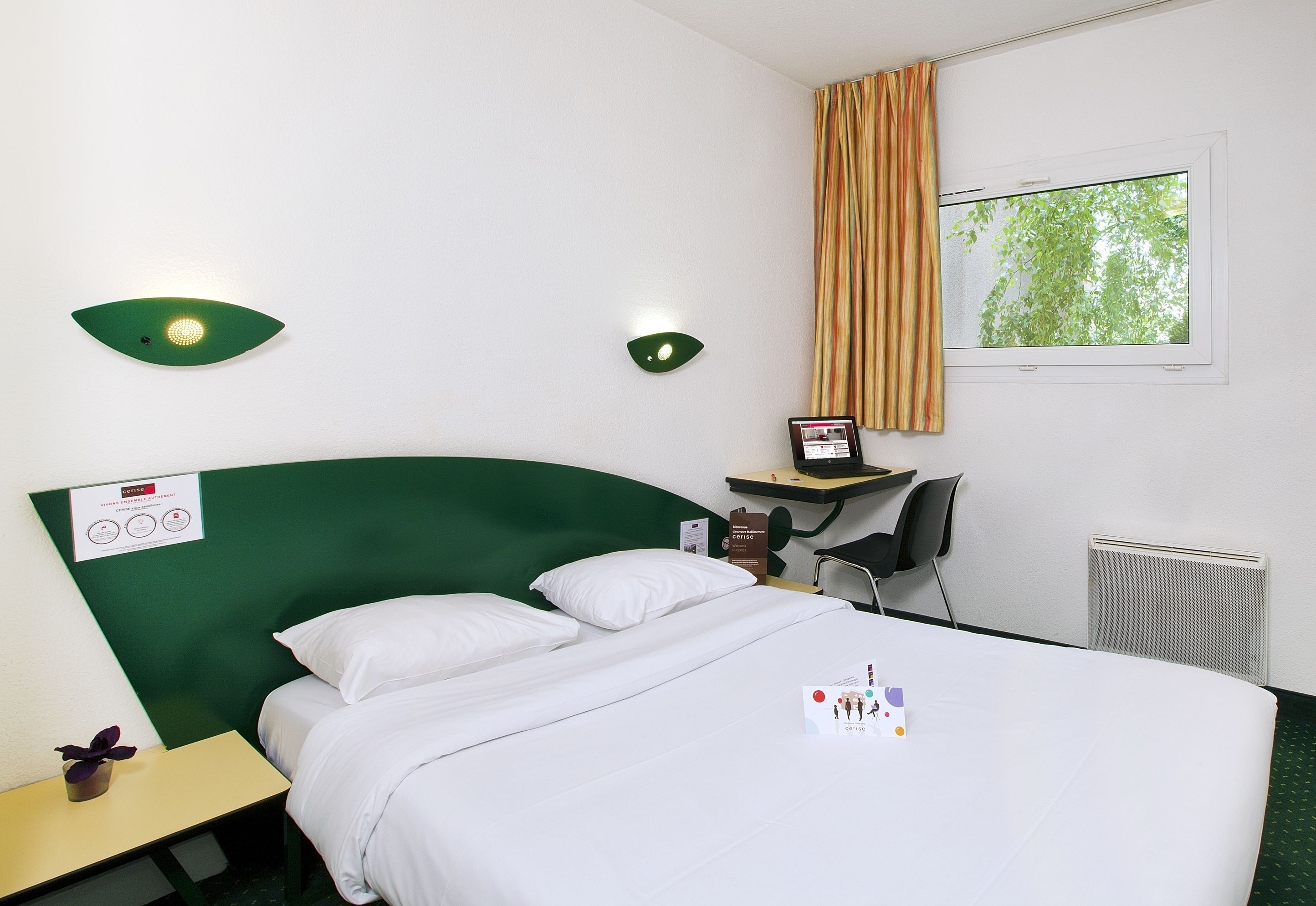A hotel room with a green headboard, white bedding, a desk with a laptop, a chair, and a window with curtains.