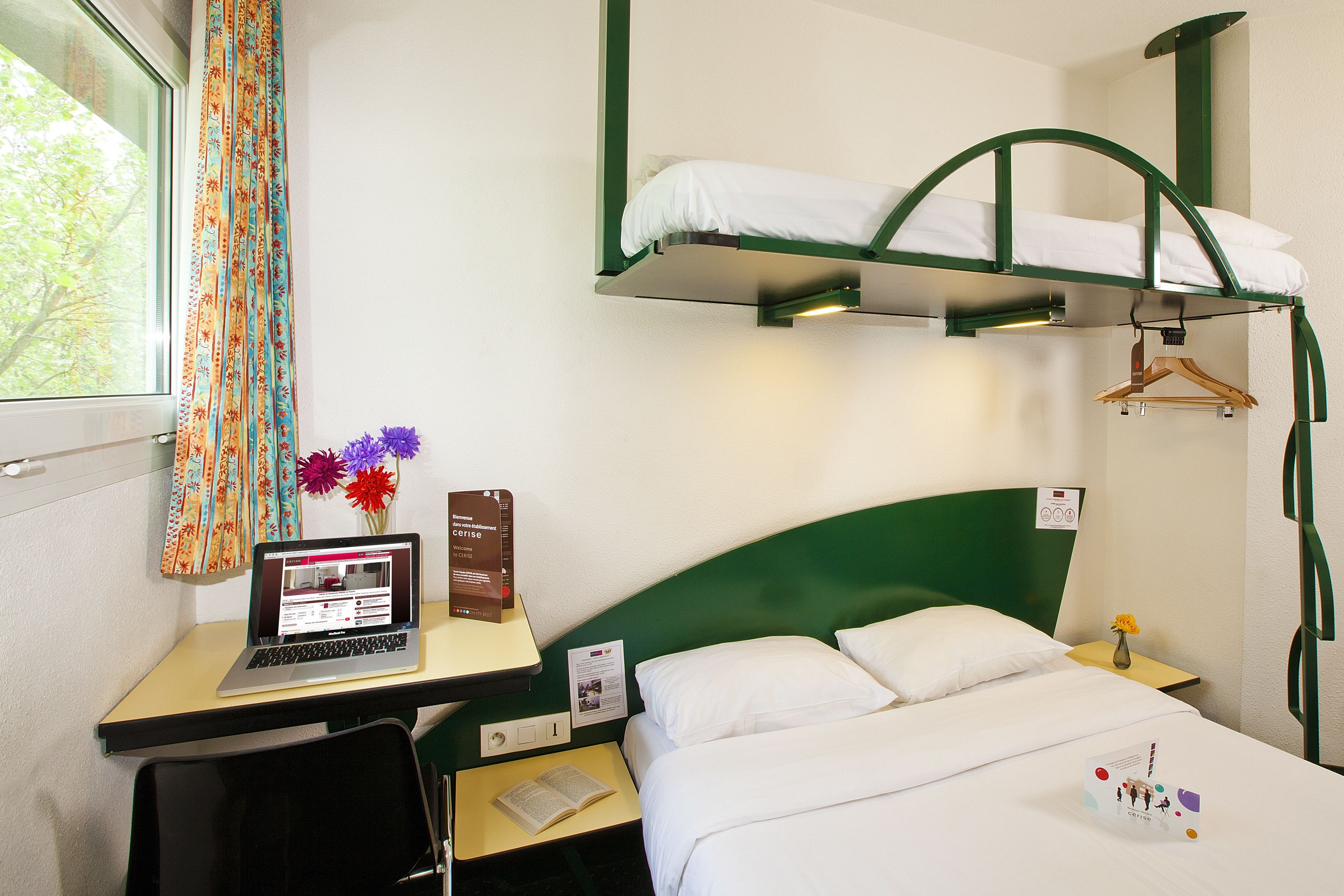 Habitación de hotel con literas, un escritorio con una computadora portátil, una silla, una mesita con un libro, un florero con flores y un perchero.