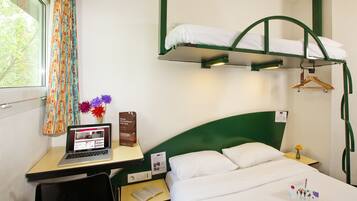 Habitación de hotel con literas, un escritorio con una computadora portátil, una silla, una mesita con un libro, un florero con flores y un perchero.
