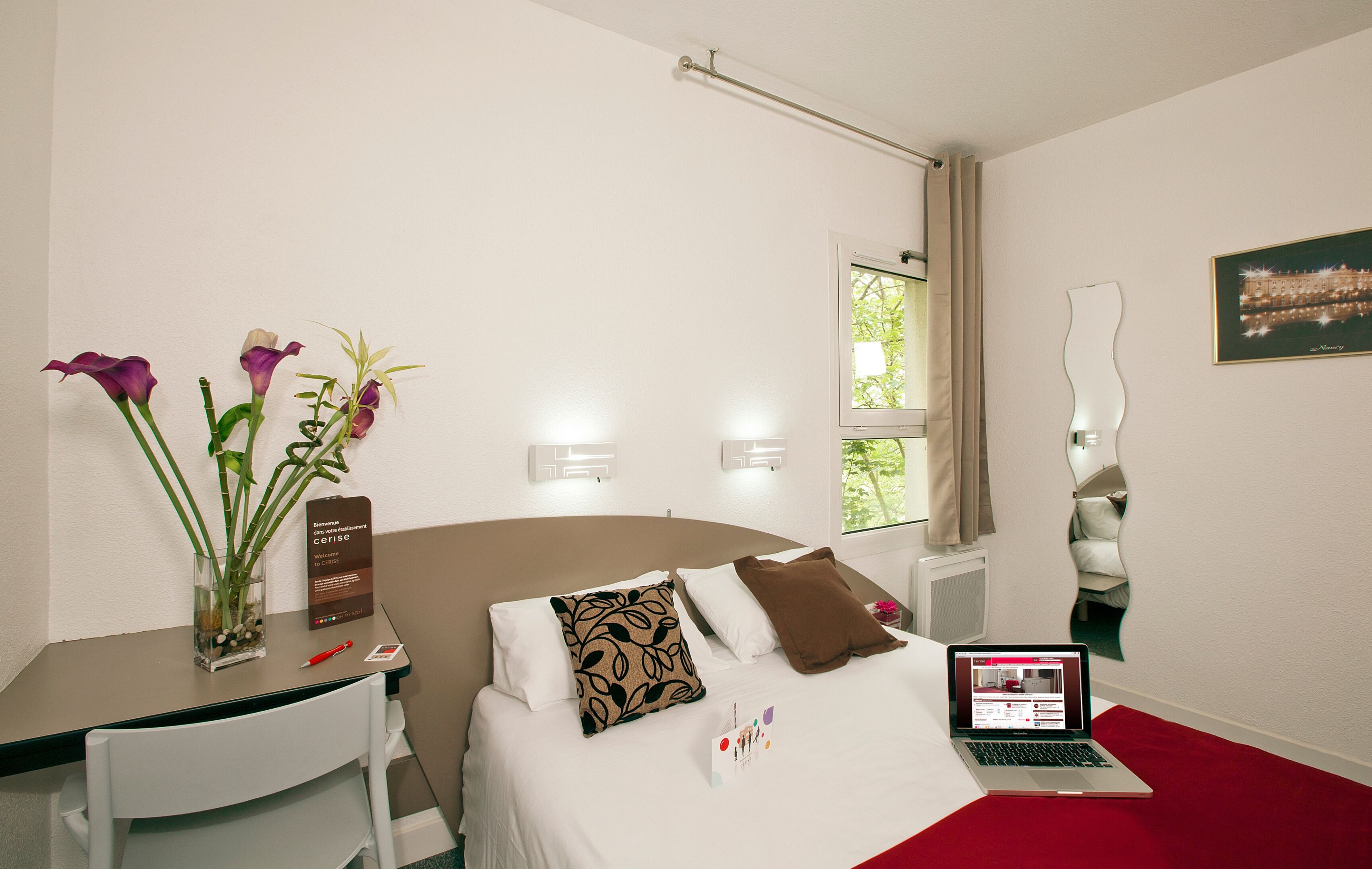 A hotel room with a bed, a desk, a laptop, and a vase with flowers.