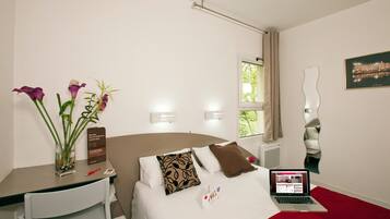 A hotel room with a bed, a desk, a laptop, and a vase with flowers.