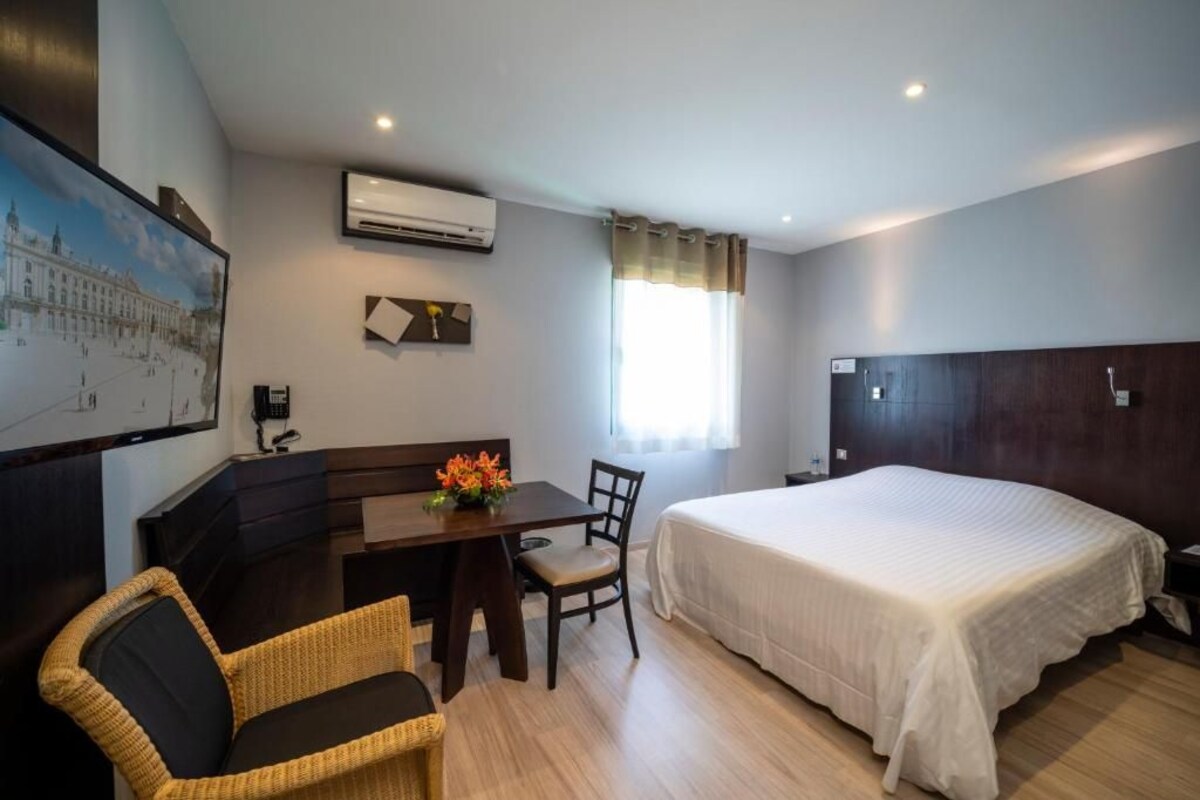A hotel room with a bed, a desk with a chair, a small table with a vase of flowers, a painting on the wall, and an air conditioning unit.