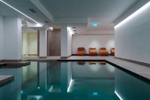Un área de piscina interior moderna con una piscina cristalina, asientos a lo largo de la pared y una señal de salida visible.