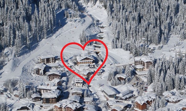 A snow-covered village with traditional chalet-style buildings clustered around a central area, surrounded by dense forest. - Dahu (Pinzolo)
