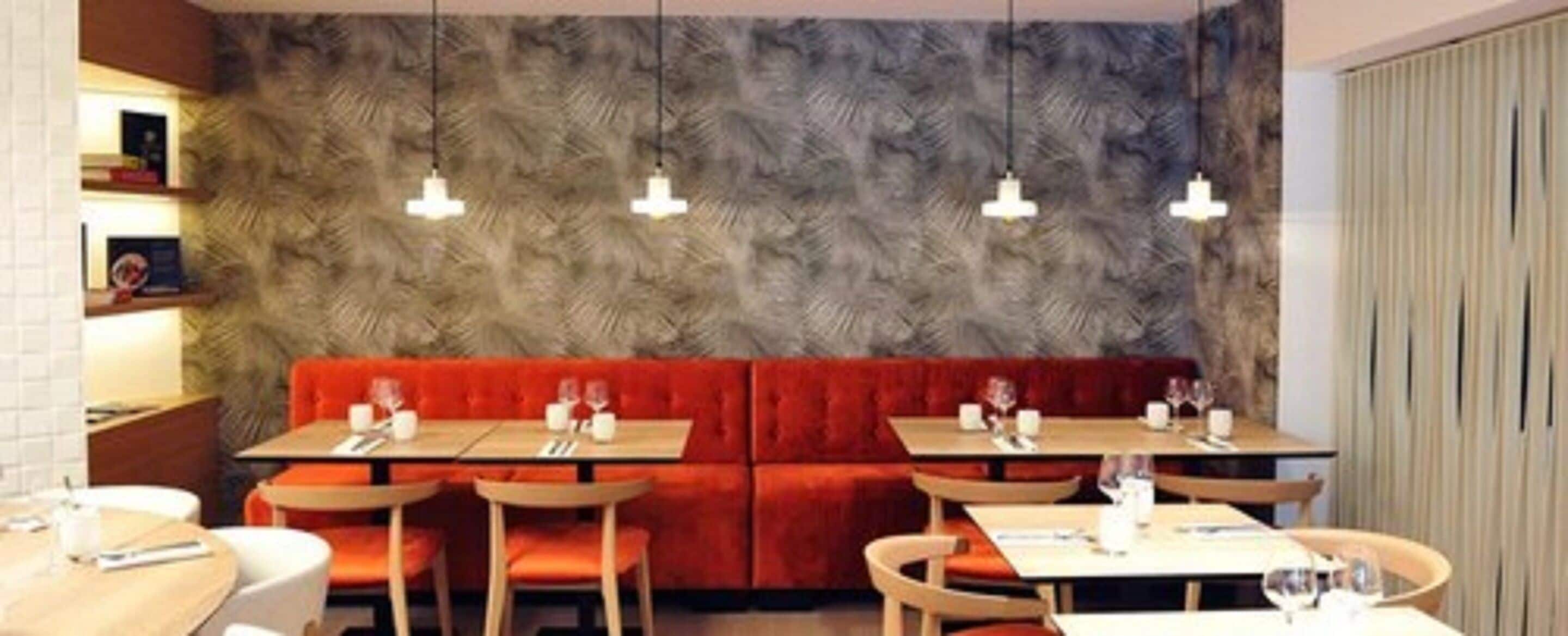 Un restaurant amb una banca vermella amb coixins, taules de fusta i lluminàries penjants.