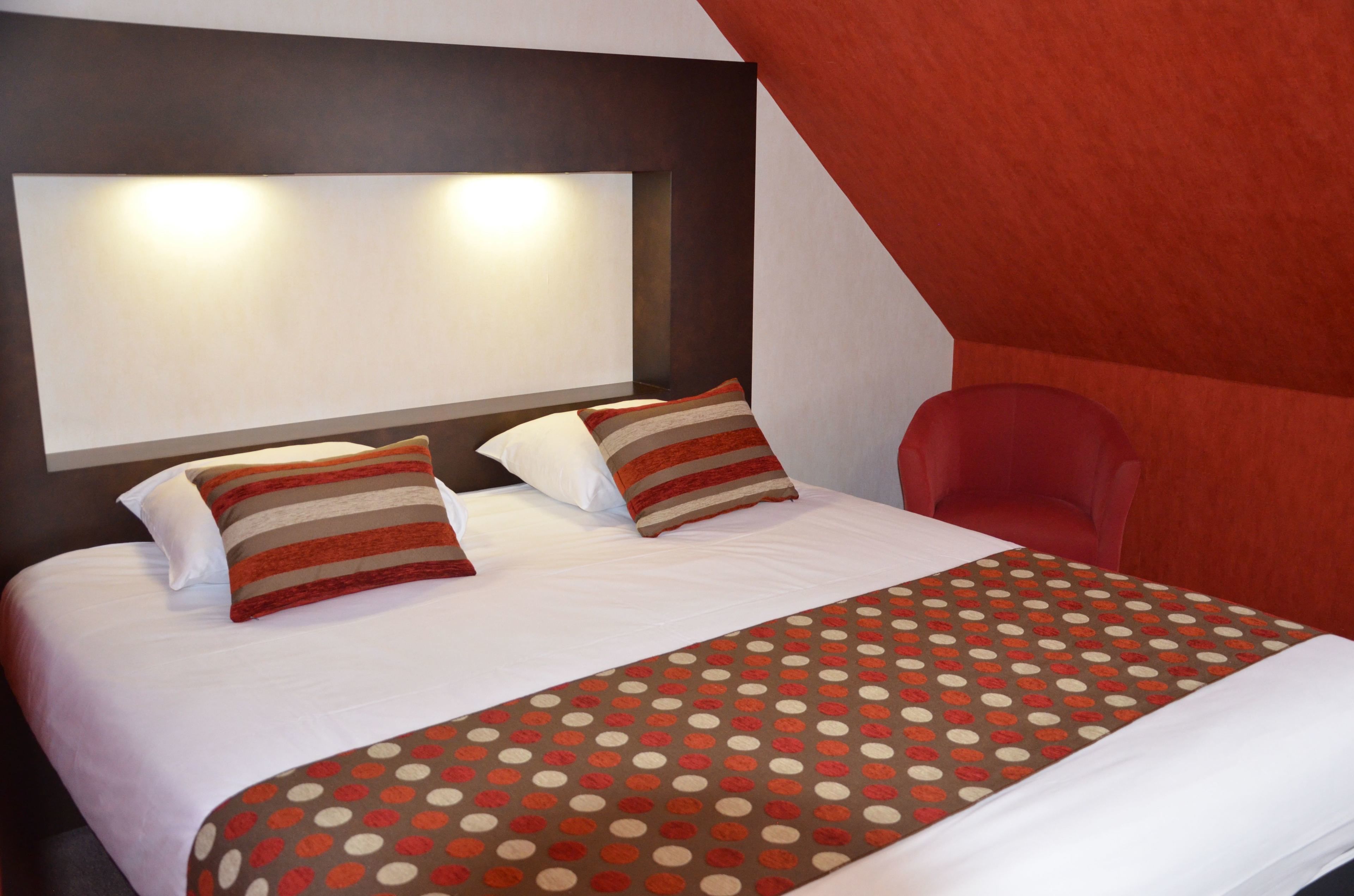 Habitación de hotel con una cama grande, dos almohadas, una manta de lunares, una silla roja y un cabecero con luces.