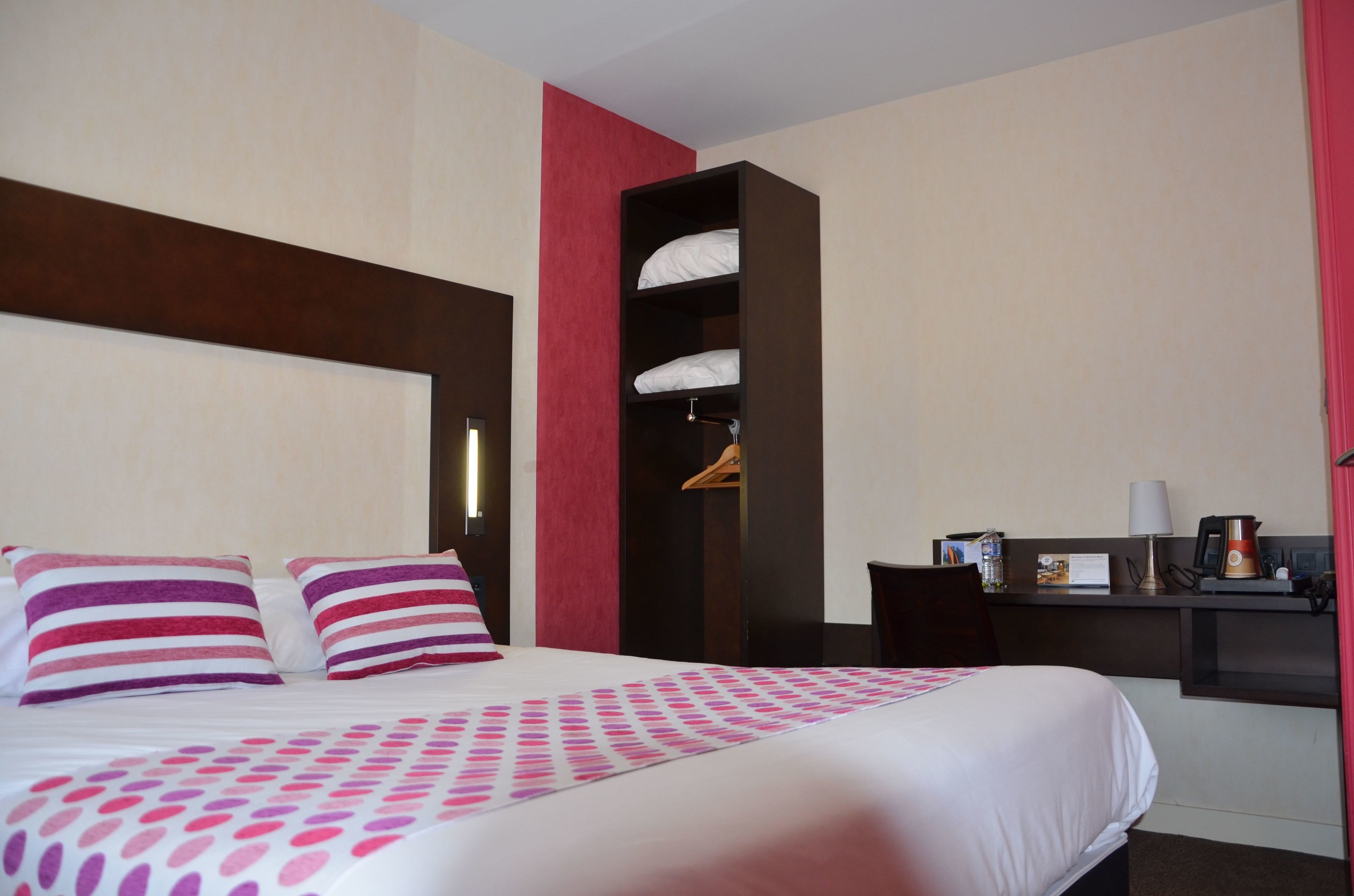 A hotel room with a large bed, a bedside table with a lamp, a small desk, and a wardrobe.
