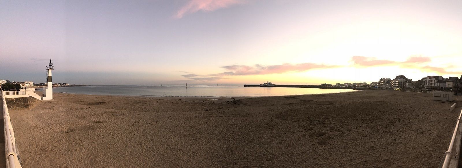 Une plage avec un phare, une mer calme et des bâtiments en arrière-plan.