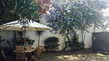 An outdoor patio area with a table and chairs under a white umbrella, surrounded by various plants and trees.