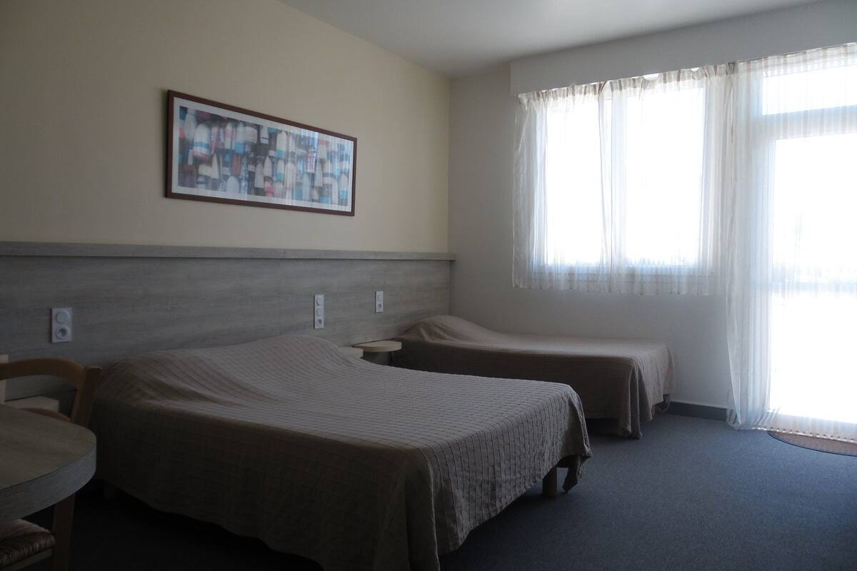 Une chambre d’hôtel avec deux lits simples, une petite table et une fenêtre avec des rideaux légers.