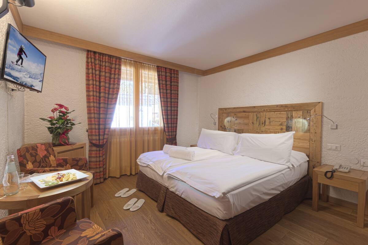 Camera d'albergo con testiera in legno, letto con lenzuola bianche, scrivania in legno con un piatto di cibo, una sedia, un tavolino con un telefono e una finestra con tende a quadri.