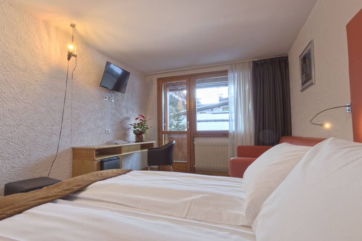 Una camera d'albergo con un letto, una scrivania con una sedia, una TV a parete e una finestra con tende.